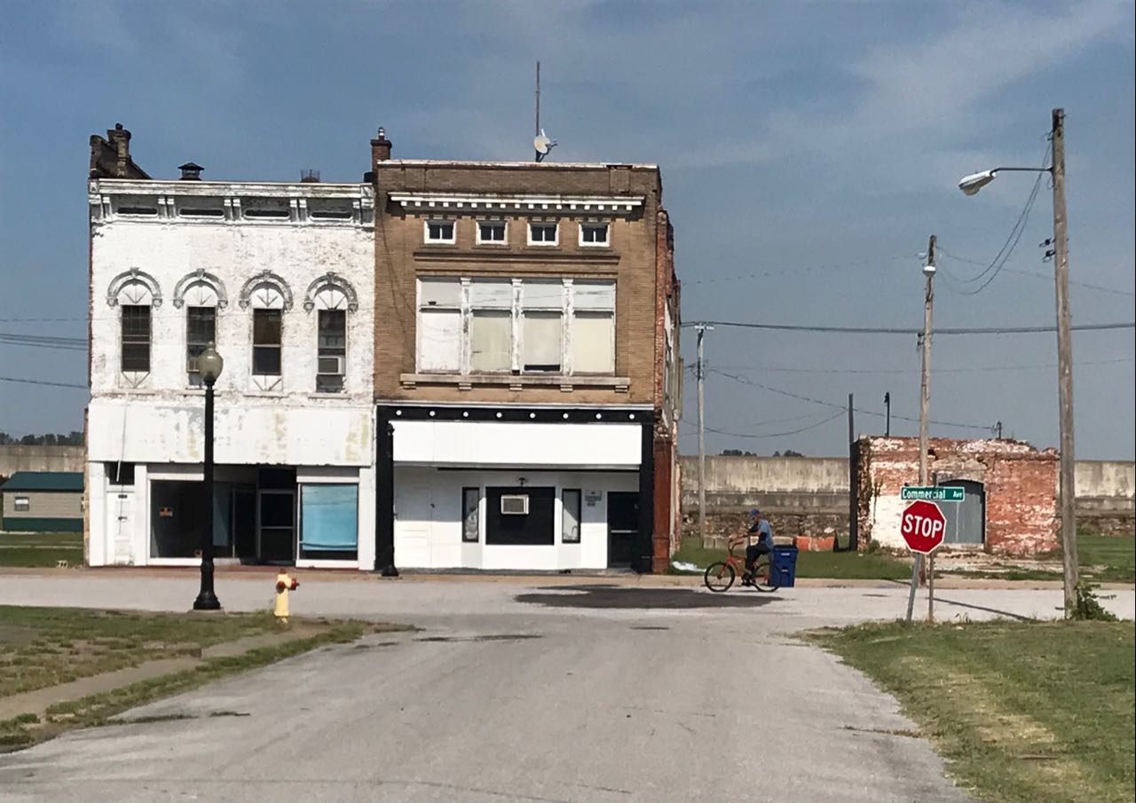 This meme is a photo of a deserted, abandoned town with crumbling buildings, a "For Sale" sign on the left, and a "For Rent" sign on the right. The text on the sign on the left reads "We have nothing. We have nothing. We have nothing." The image is a humorous commentary on the state of the town's economy and the lack of opportunities or resources available to its residents.
